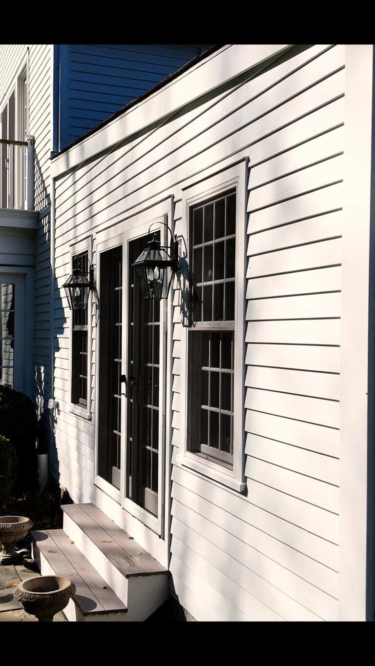 Home addition builder Omega Development in Connecticut. White house exterior with two black‑framed glass doors centered between large multi‑pane windows, flanked by black lantern lights and two stone planters in the foreground.