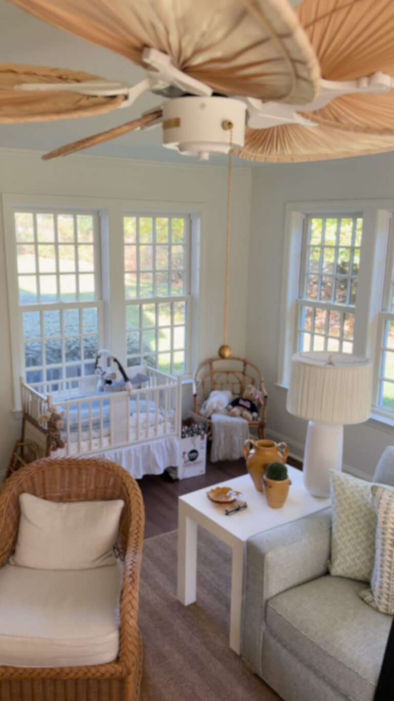 Home addition contractor in CT, Omega Development. Bright, cozy nursery or sitting room with a white crib, rattan chair, gray couch, wicker armchair, large windows, and a ceiling fan with leaf‑shaped blades.