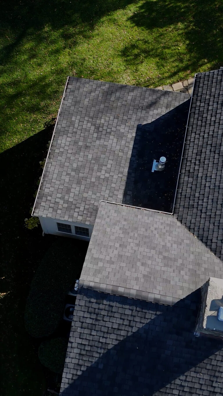 Home addition contractor Omega Development. Aerial view of a gray‑shingled residential roof with multiple intersecting slopes and a vent pipe, surrounded by a neatly maintained lawn and tree shadows.
