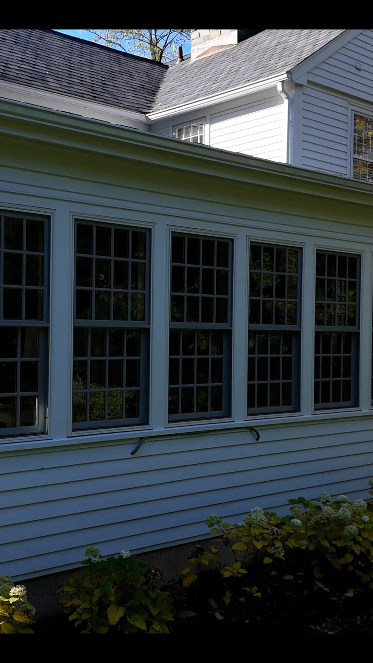 Home addition contractor Omega Development, Connecticut. Exterior wall of a white house with large grid‑pane windows, dark frames, gray shingle accents above, metal brackets mounted below the windows, and flowering plants in front.