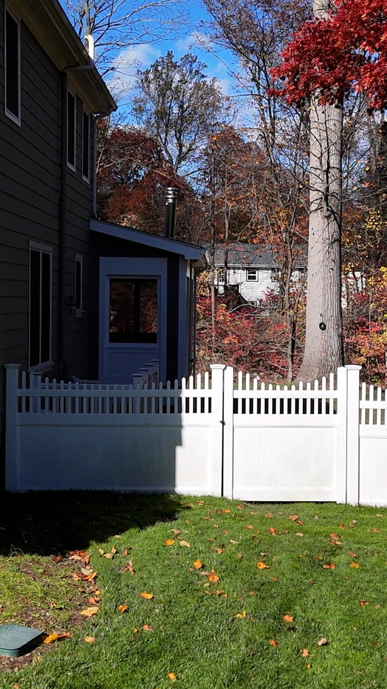 Home addition builder in Connecticut Omega Development. Backyard scene with a white picket fence and gate, fallen autumn leaves on the grass, a dark‑colored house behind the fence, and a tall red‑leaved tree to the right under a clear blue sky.