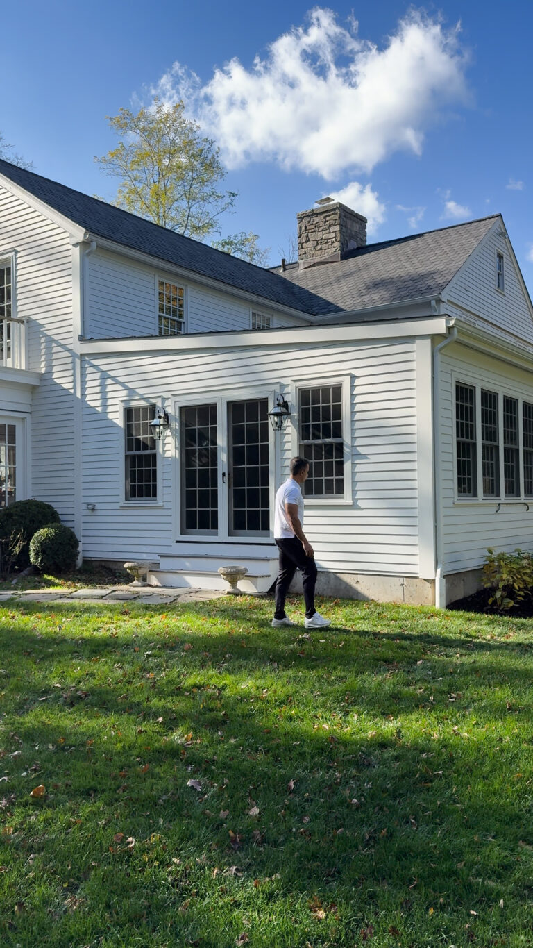 Home addition contractor Omega Development. White house with large grid‑pane windows and a stone chimney, viewed from the lawn as a person walks across the grass on a bright, sunny day.