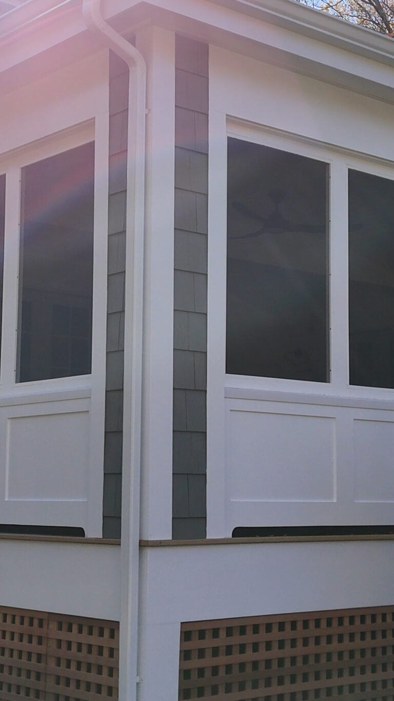 Home addition contractor in CT, Omega Development. Corner view of a screened‑in porch with white trim, gray shingle accents between large dark windows, a ceiling fan inside, and a wooden lattice skirt below.