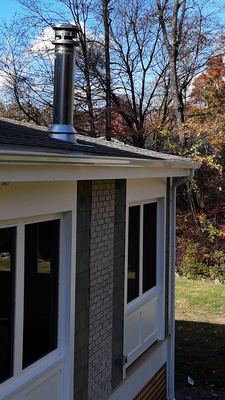 Home addition contractor, Omega Development. Exterior wall of a house with a metal chimney pipe extending above the roof, large windows below, and mixed siding materials including white trim, gray shingles, and a vertical brick accent.