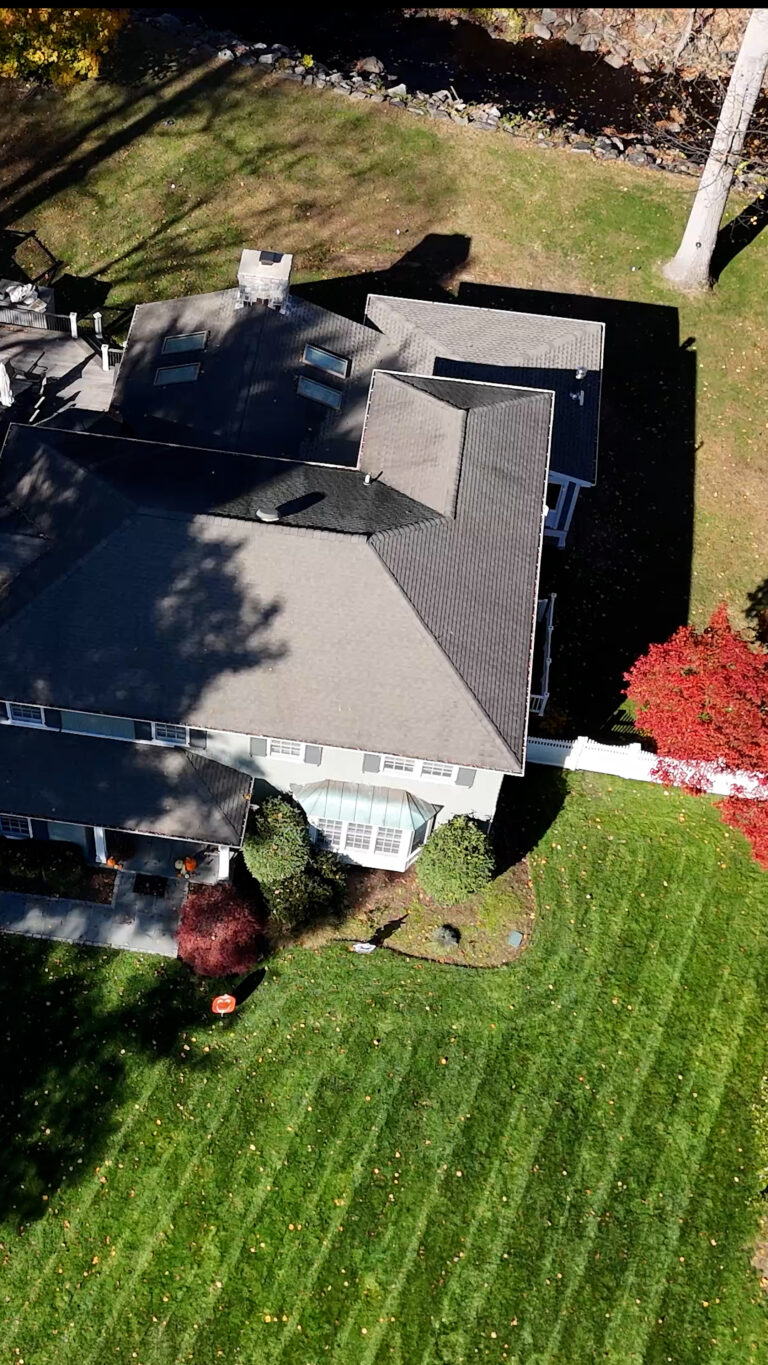 Home addition contractor in Connecticut, Omega Development. Aerial view of a large house with a complex roof, surrounded by a neatly striped lawn, trees, shrubs, and a small stream along the edge of the property.