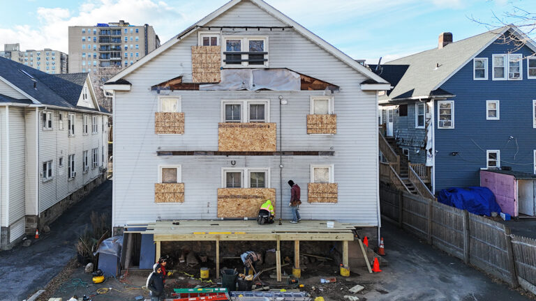 Deck installation contractor in Connecticut, Omega Development. Multi‑story white house under renovation, with boarded‑up windows, workers on a newly built wooden deck, and construction tools and materials around the site.