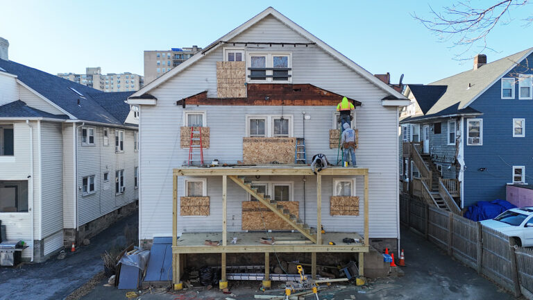 Deck installation contractor Omega Development. Multi‑story white house under renovation with boarded‑up windows, a new wooden porch structure being built, and two construction workers on the upper level.