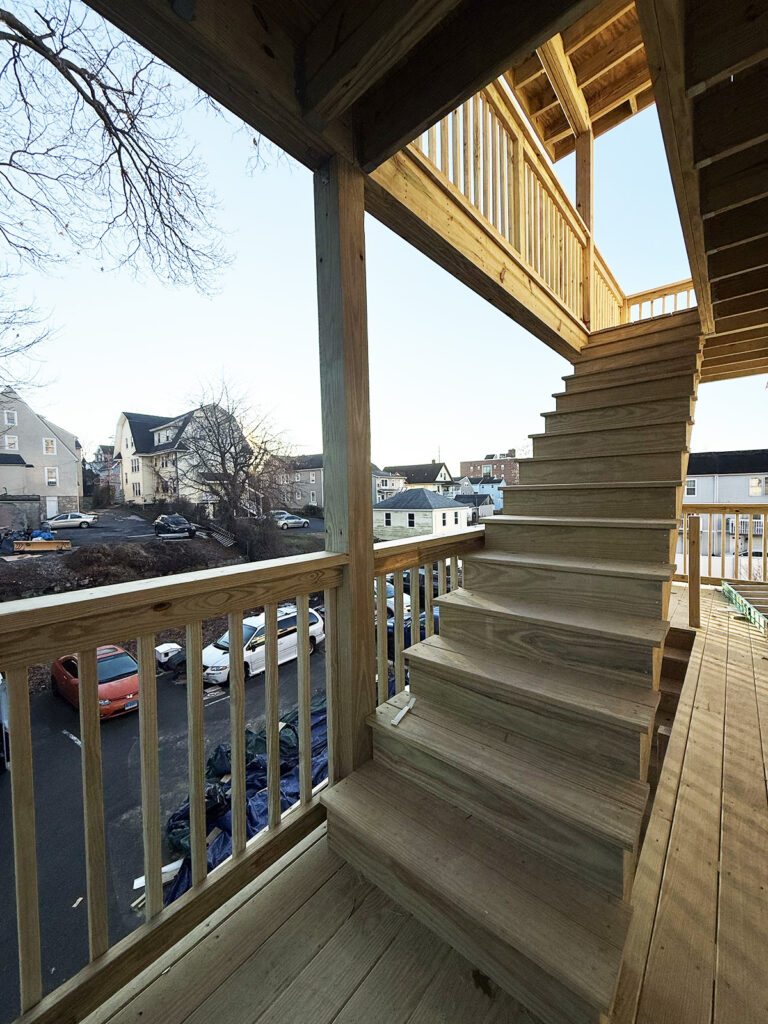 Deck construction in Connecticut by Omega Development. Outdoor wooden staircase leading to an upper covered landing, built from unfinished lumber and attached to a multi‑level building, with parked cars and neighboring houses in the background.