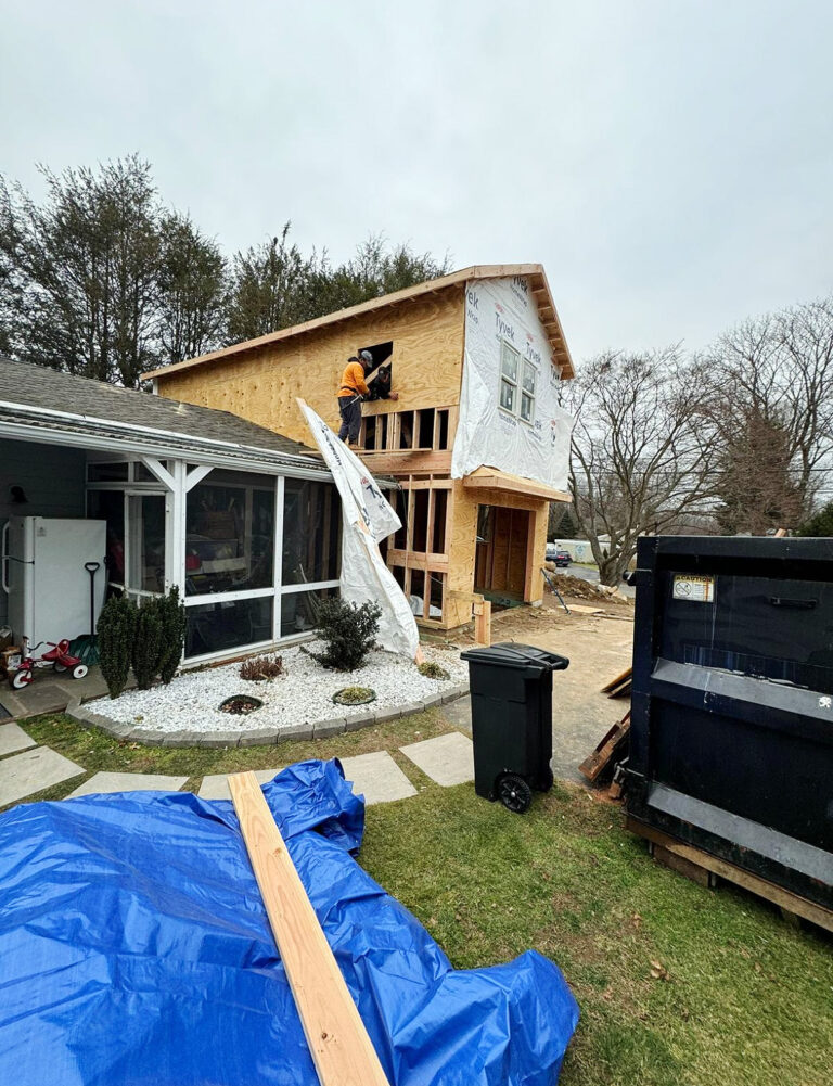 Contractor for new construction in Connecticut, Westport, Omega Development. Exterior of a white house with large grid‑pane windows, dark frames, gray shingle accents above, metal brackets mounted below the windows, and flowering plants in front.