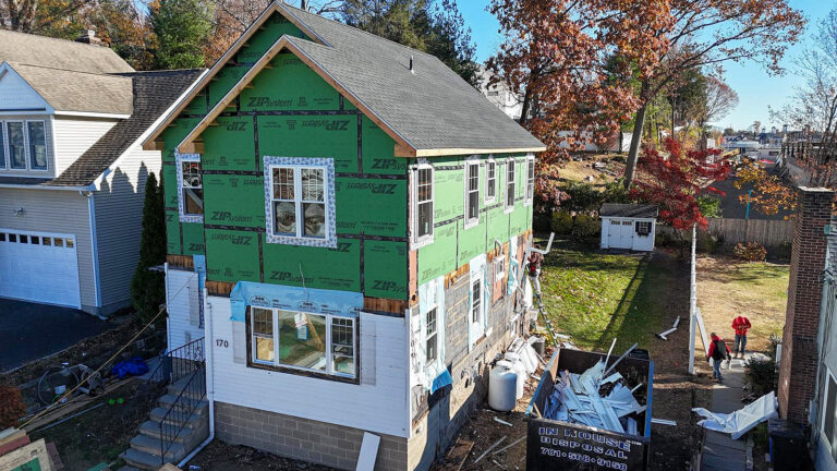 Contractor for new construction in CT by Omega Development. House exterior under renovation with green sheathing on the upper level, partially installed white siding below, exposed concrete blocks, a large debris dumpster in front, and two people walking past on the sidewalk.