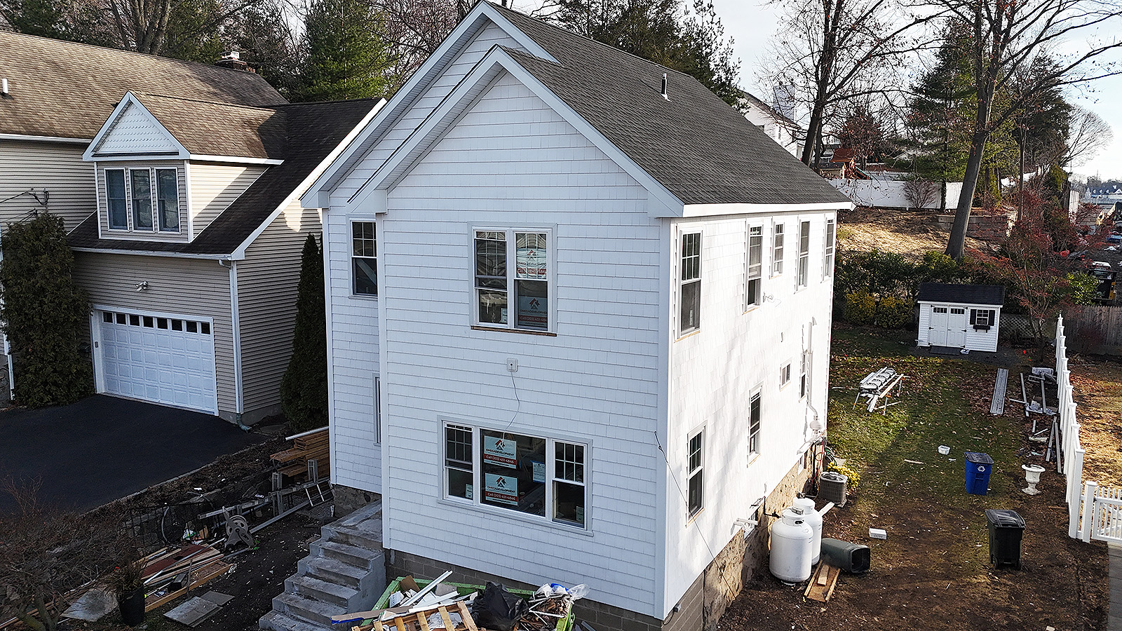 Home remodeling services in Connecticut by Omega Development. White two‑story house with new siding and windows, construction materials scattered in the yard, and a beige neighboring house beside it.