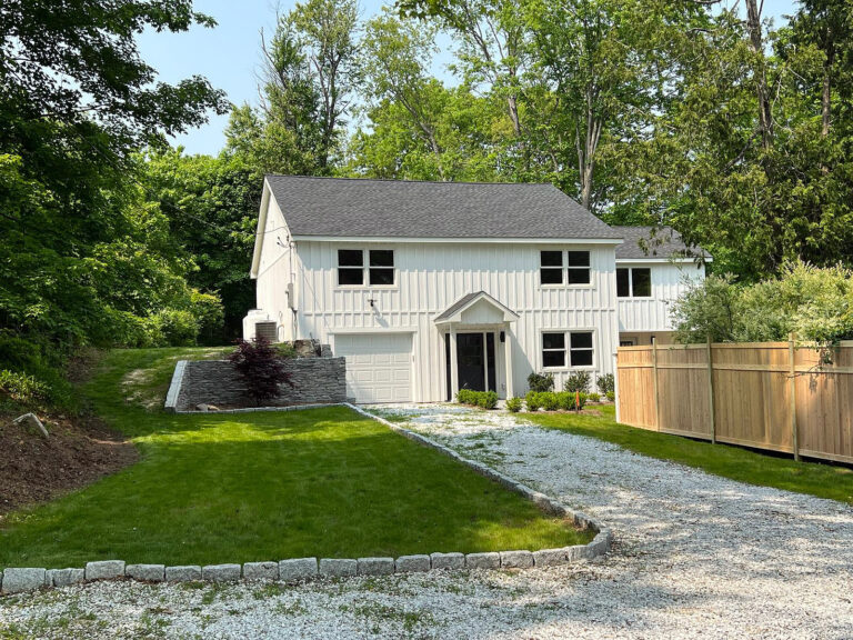 Contractor for new construction, Omega Development CT. Two‑story white house with a dark roof, gravel driveway, and surrounding trees.