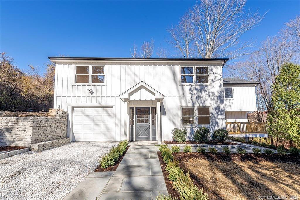 Home renovaton in Connecticut. Two‑story white house with vertical siding, a central gabled entry, a single‑car garage, gravel driveway, stone retaining wall, and landscaped front yard.