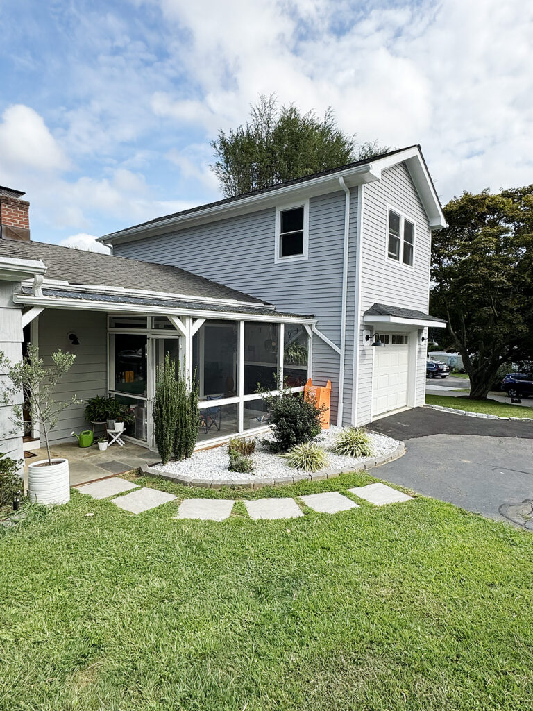 Driveway paving contractor in Connecticut. Two‑story house with light gray siding, a screened‑in porch, landscaped front garden with white gravel and shrubs, and a curved stone path leading across the lawn.
