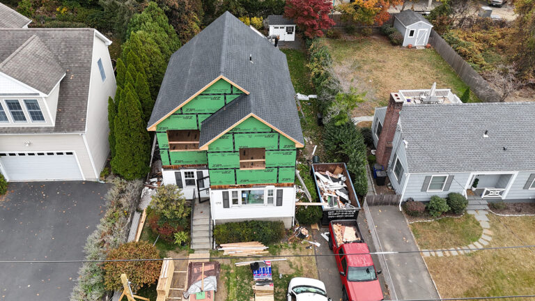 Contractor for new construction in Westport CT Omega Development. Aerial view of a house undergoing renovation, with a newly added second story covered in green sheathing, construction materials in the yard, a red pickup in the driveway, and a large dumpster beside the house.