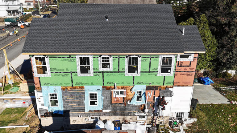 Contractor for new construction in Westport, Connecticut, Omega Development. House exterior under renovation with green and brown sheathing panels, partially installed gray shingles, a worker on a ladder applying siding, and construction materials scattered around the yard while roadwork takes place nearby.