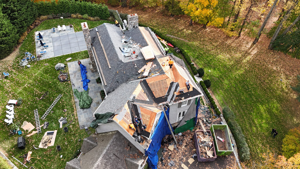 Roofing installation and repair by Omega Development LLC in Westport CT and beyond. Aerial view of a house undergoing major roof renovation, with workers removing old materials and installing new wooden panels, tarps covering parts of the structure, tools and ladders around the yard, and a large green dumpster filled with debris.