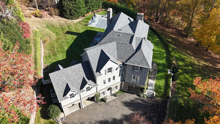 Roofing contractor in Westport, Connecticut, Omega Development. Aerial view of a large multi‑story home with a complex roof, stone and siding exterior, a paved driveway, and a backyard patio with outdoor furniture, surrounded by a manicured lawn and autumn trees.