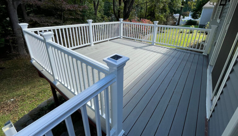Deck construction by Omega Development. Elevated backyard deck with gray composite flooring and white railings, featuring decorative post caps and supported by wooden posts.