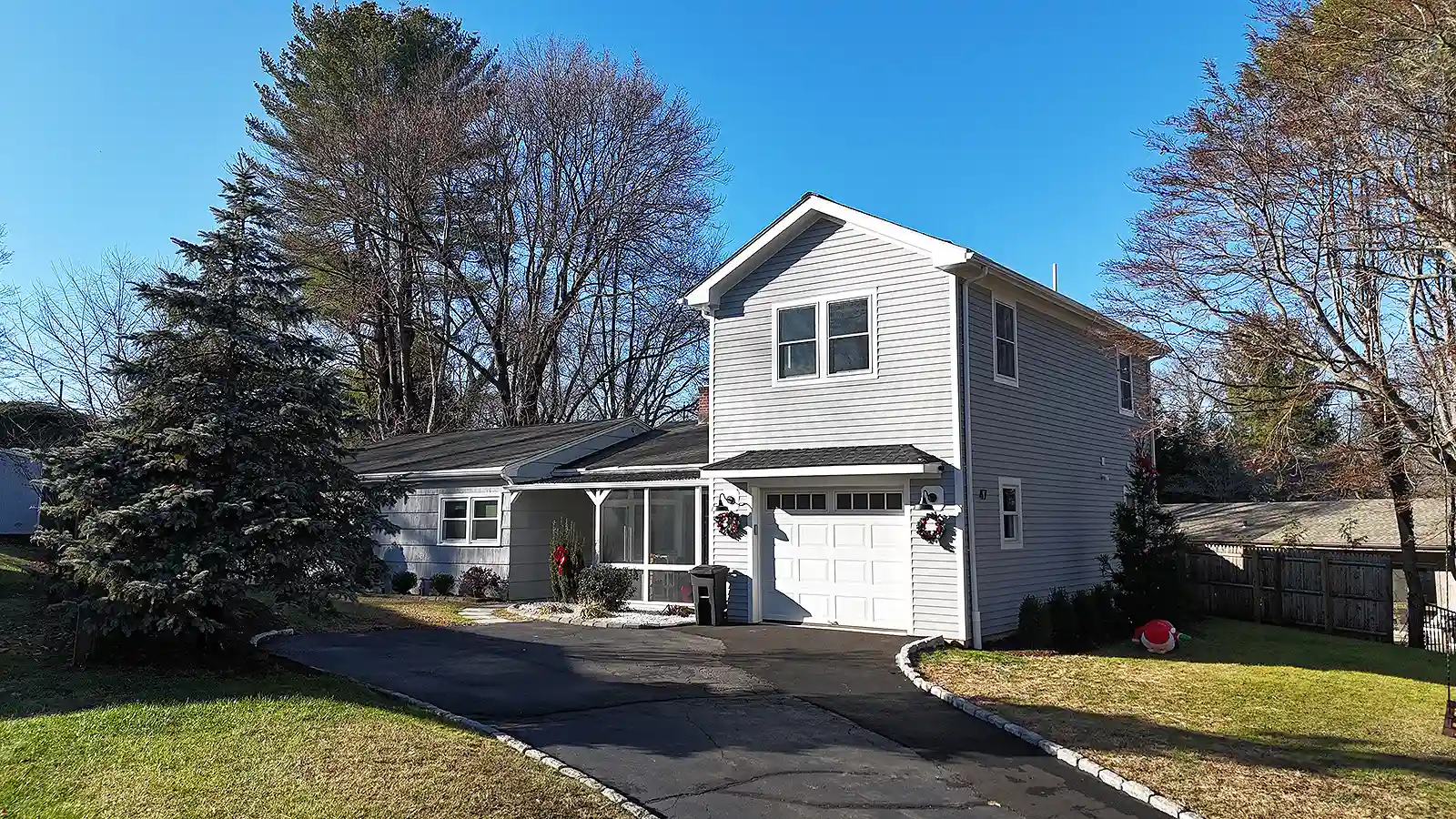 Driveway paving contractor in CT. Two‑story gray house with a garage, green lawn, and holiday decorations in the yard.