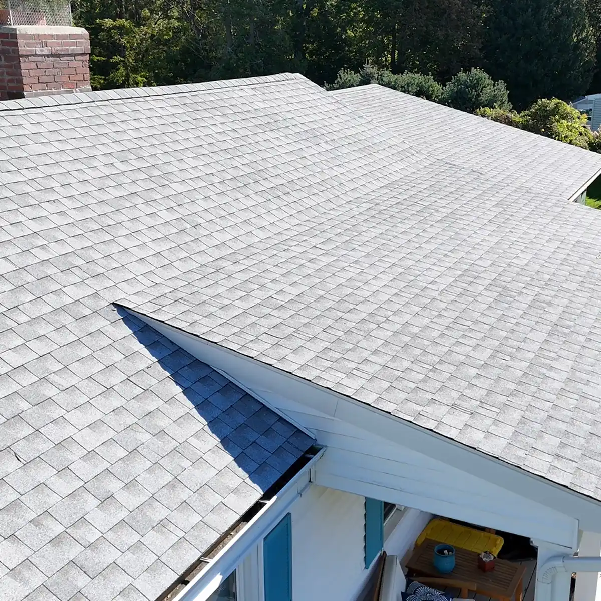 Roofing contractor Omega Development LLC Westport, Connecticut, residential roof with light gray asphalt shingles, multiple rooflines, a brick chimney, and surrounding greenery visible from an elevated angle.
